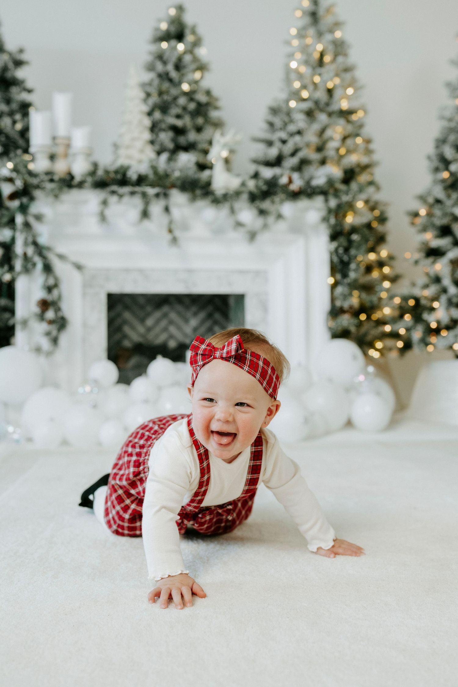 Baby wearing red plaid outfit crawls and smiles on white floor with decorated Christmas trees in background.