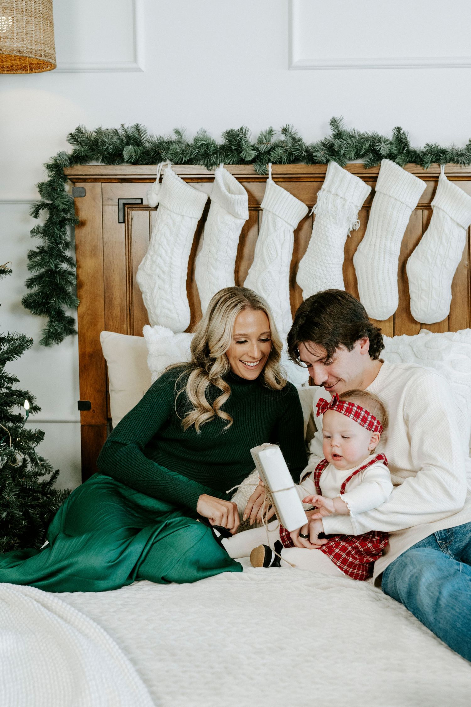Family cuddles together on white bedding with Christmas stockings and greenery decorations hanging above.