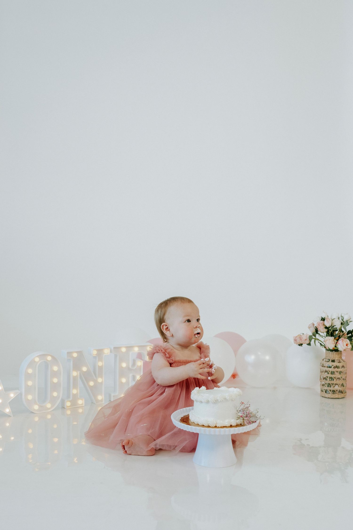 Baby in pink tulle dress crawls towards white birthday cake with illuminated 'ONE' sign in background.