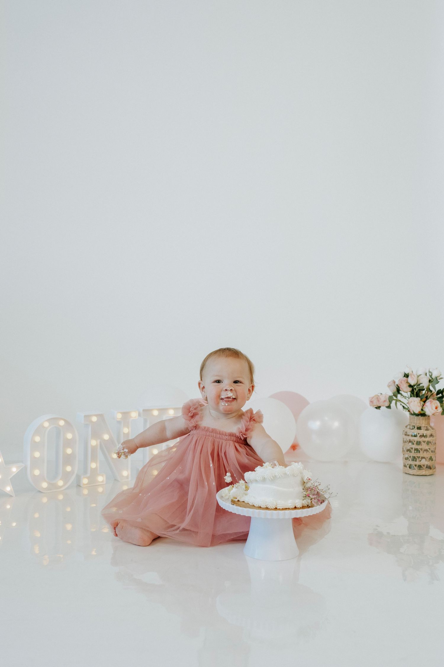 Baby in pink dress reaches for white birthday cake during first birthday photo shoot with lit marquee letters.