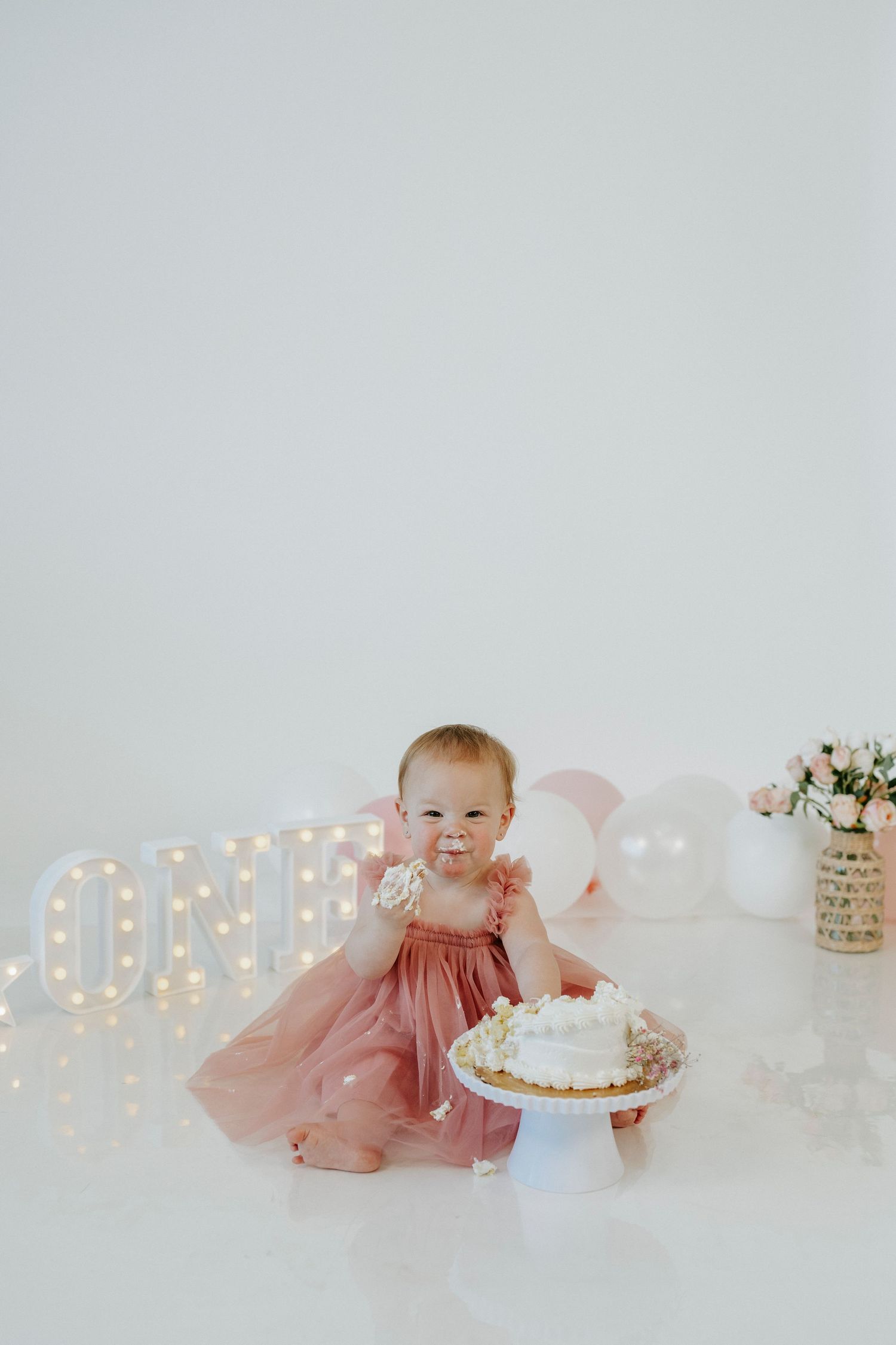 A baby in a pink dress crawls on a white backdrop with illuminated letters and a cake for a first birthday photoshoot.
