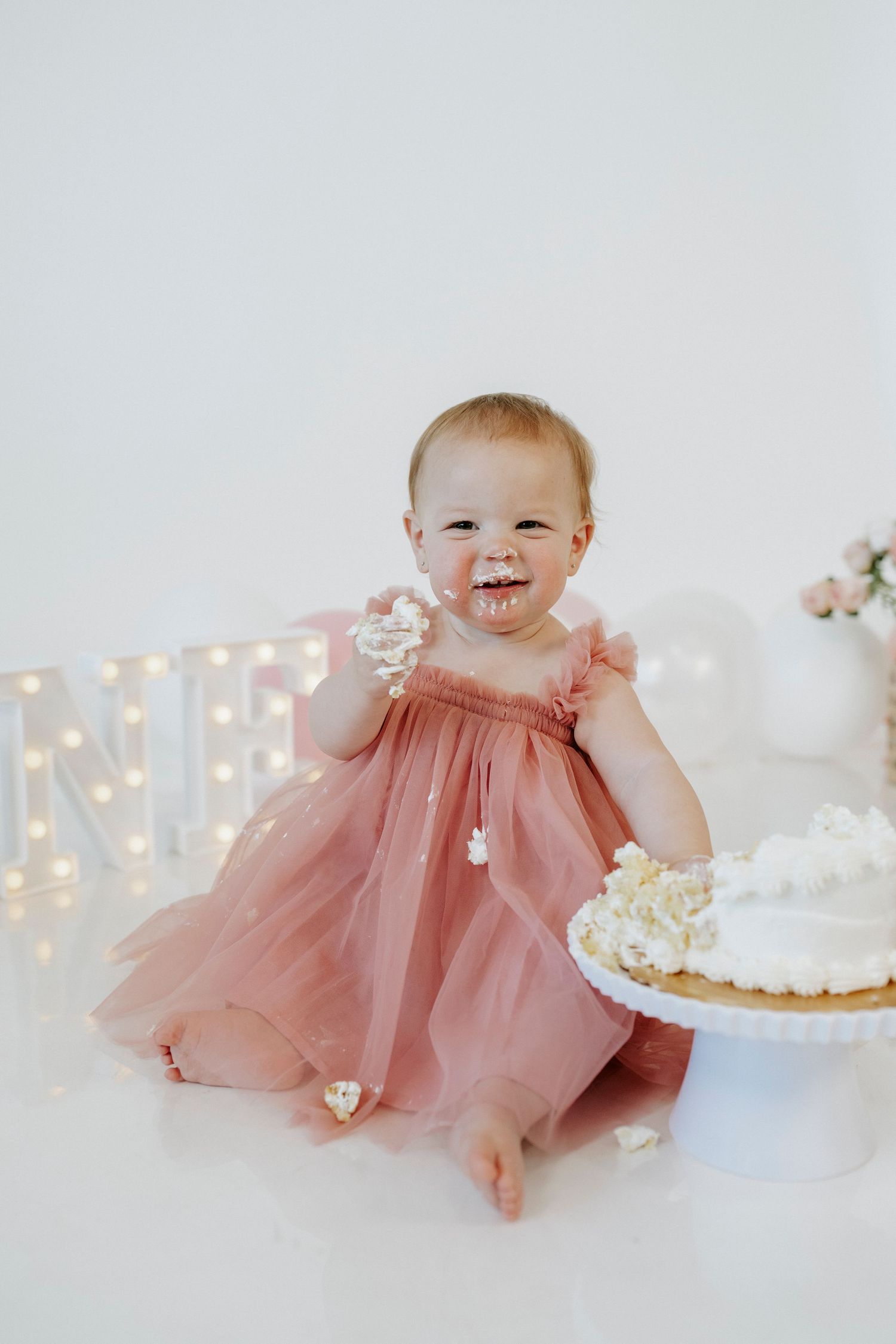 Baby in pink tulle dress smiles during cake smash photo session with twinkling lights in background.