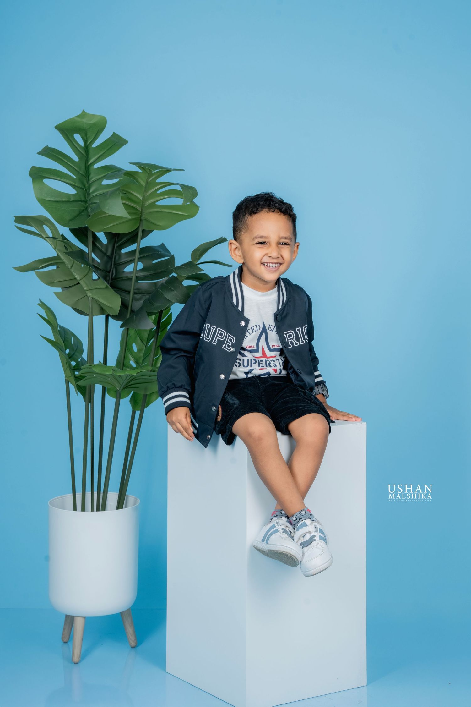 Stylish photo series showing a child in a black jacket on a white cube against a blue backdrop with a monstera plant.