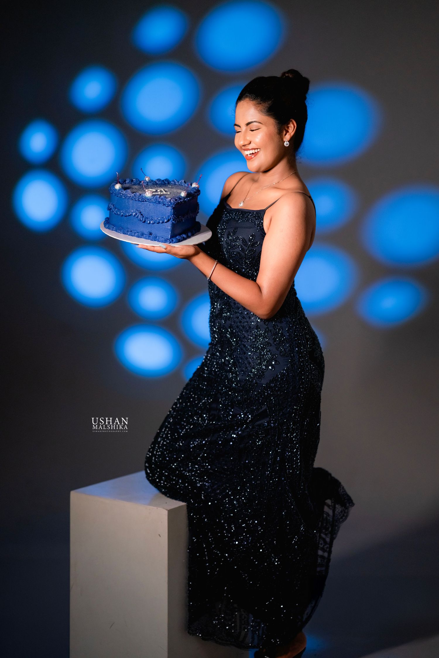 A person in a sparkling black dress poses against a dark backdrop with blue circular light patterns.