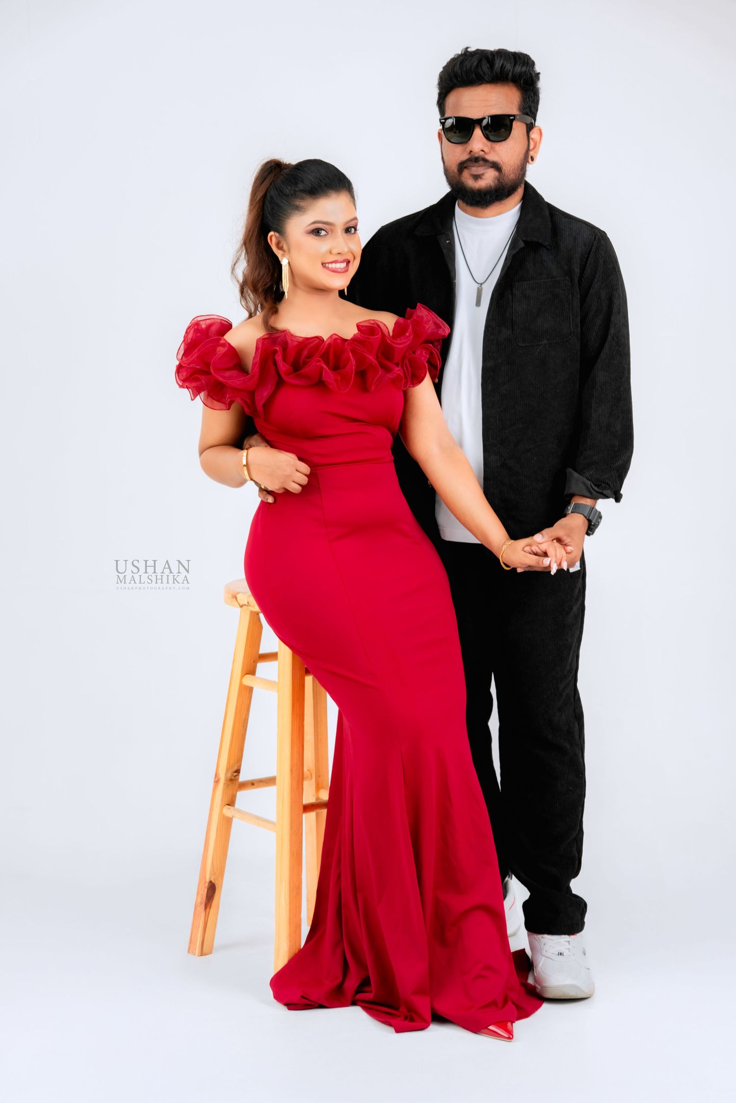 A couple poses together in a studio setting with one person wearing a red dress and the other in black attire.