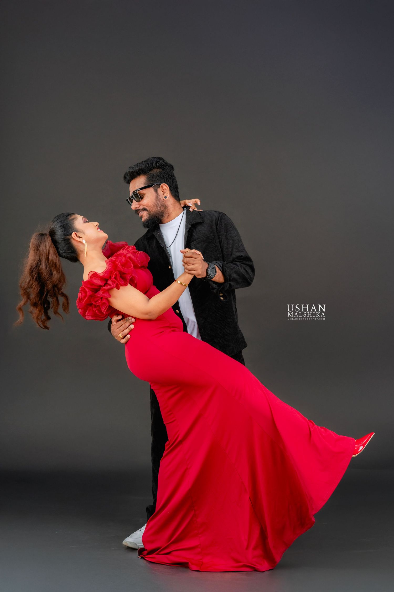 A couple performs a romantic dance dip pose, with the woman wearing a flowing red dress against a dark studio backdrop.