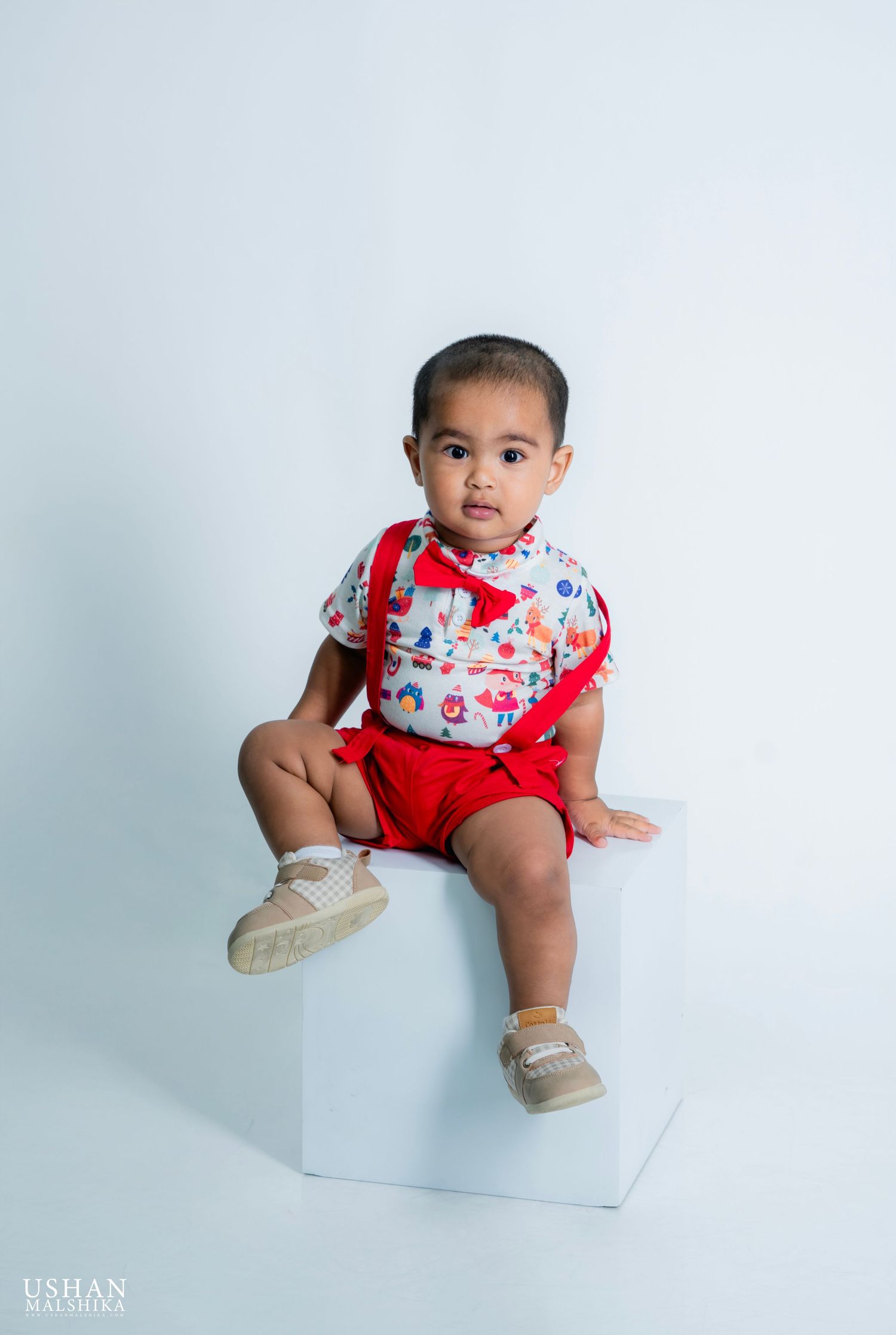 A baby in red and white floral outfit sits on a white cube against a light blue studio backdrop.