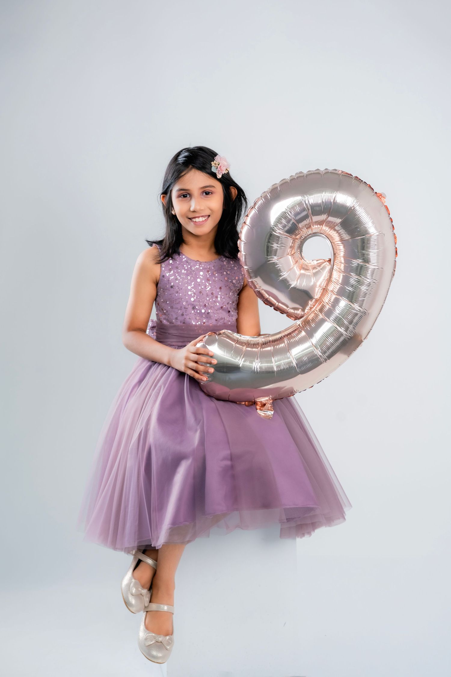 A young girl in a purple tulle dress poses with a large rose gold number 2 balloon in a photo series.