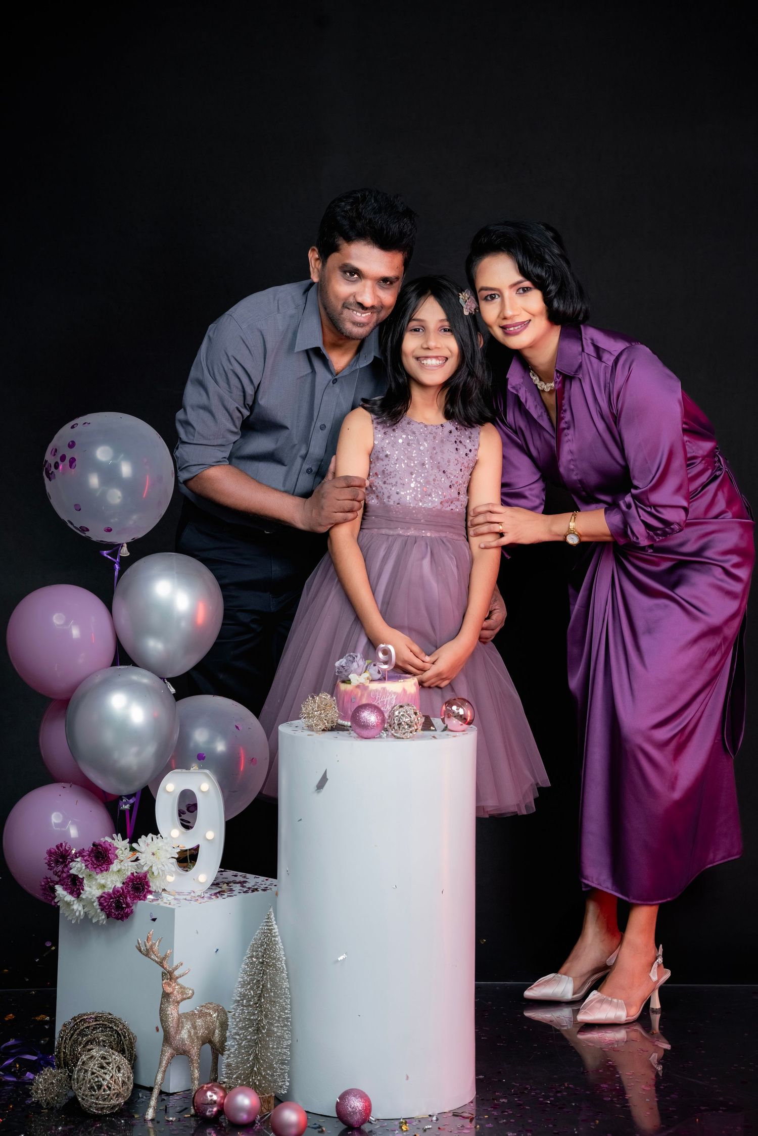 A couple poses with a white cake and purple and silver balloons during a pregnancy announcement photoshoot.