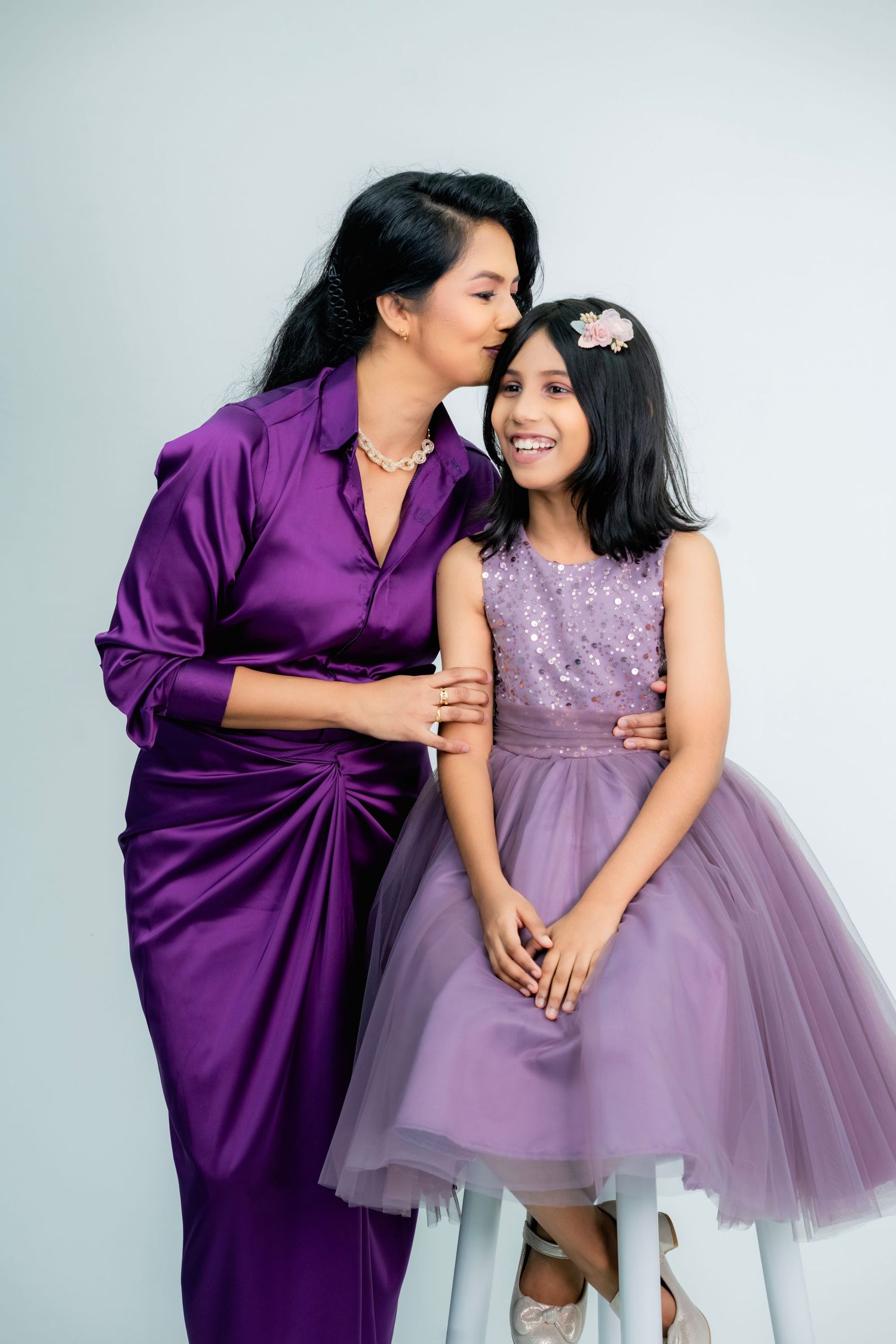 Two people wearing coordinating purple outfits share a tender moment against a light gray studio backdrop.