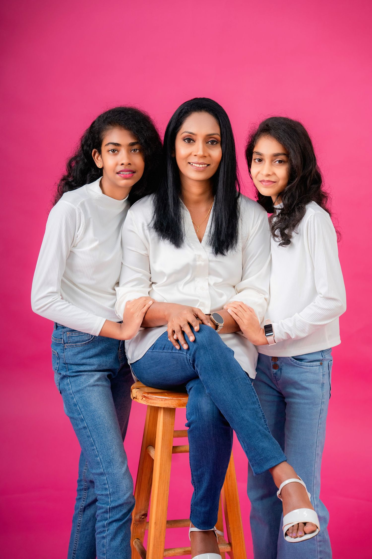 Three people in white tops and blue jeans pose together against a bright pink background.