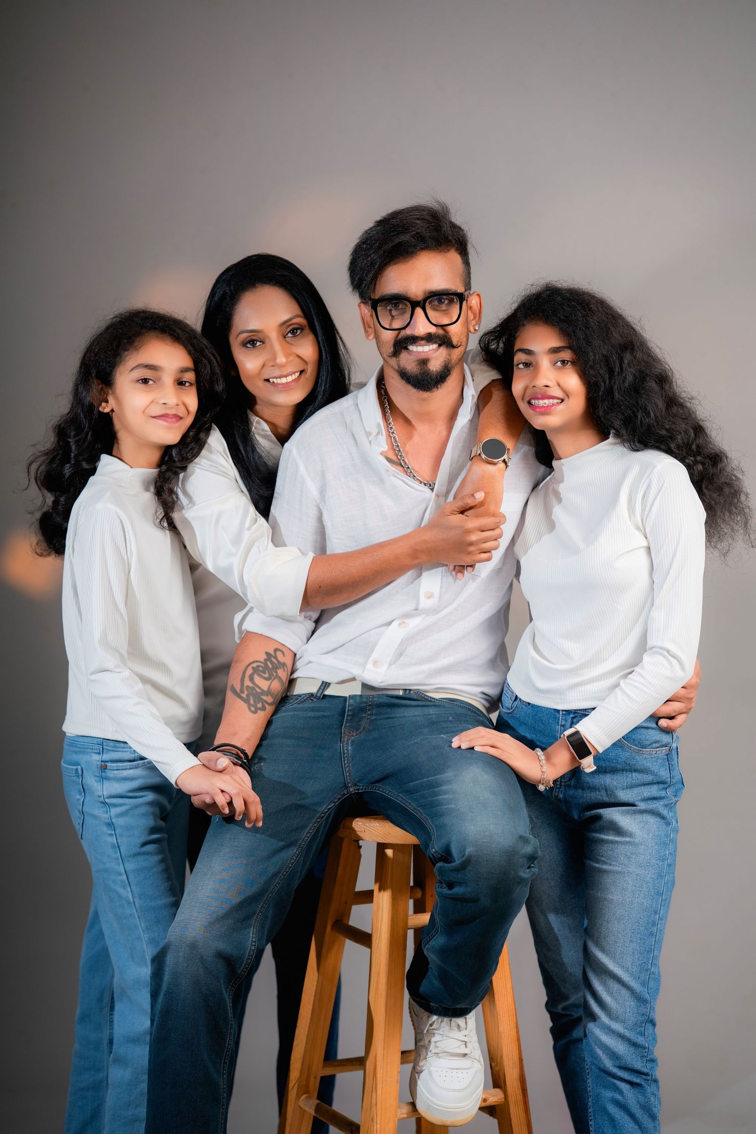 Family wearing coordinated white shirts and blue jeans poses playfully together on wooden stool against gray backdrop.