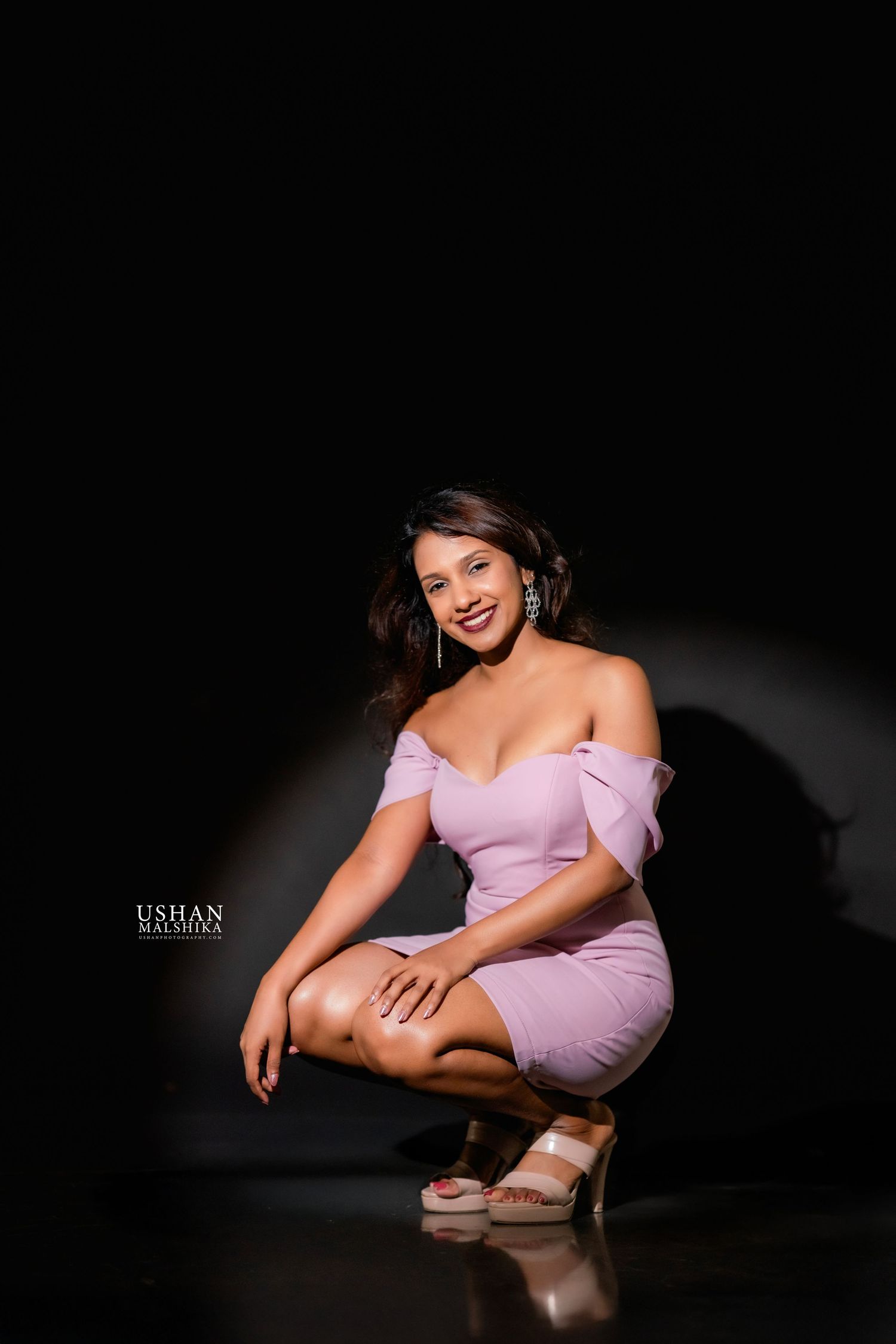 A figure in a soft pink off-shoulder dress poses elegantly in a dramatic low-light studio setting.