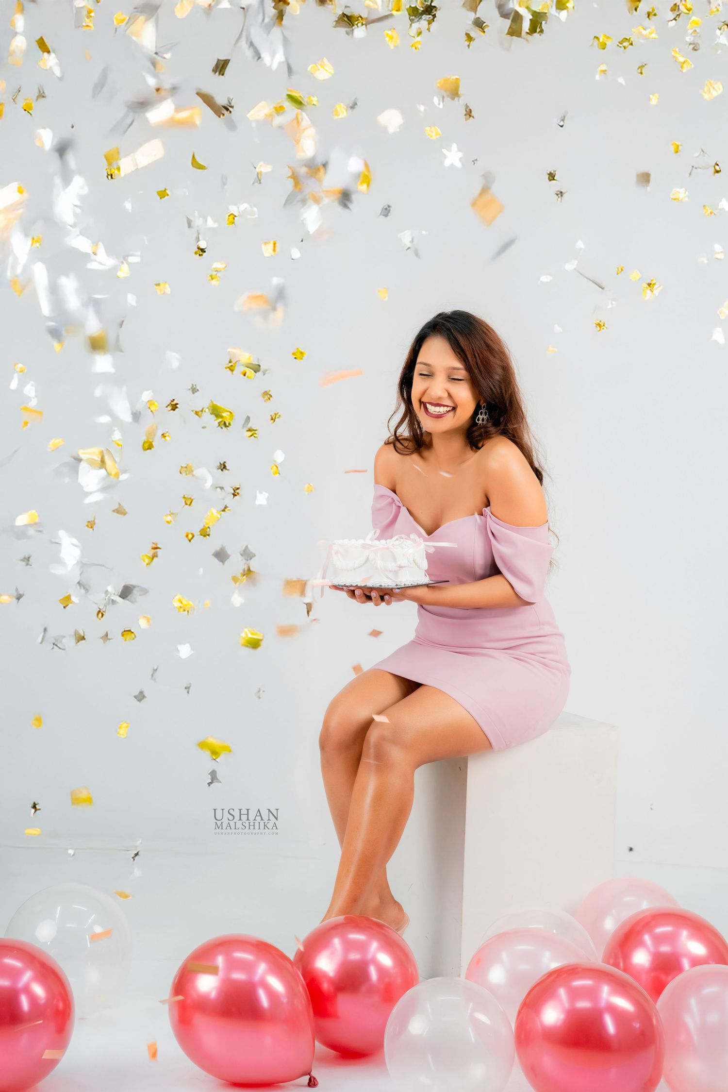 Woman in pink dress celebrates with gold confetti and pink balloons against light gray backdrop in studio setting.