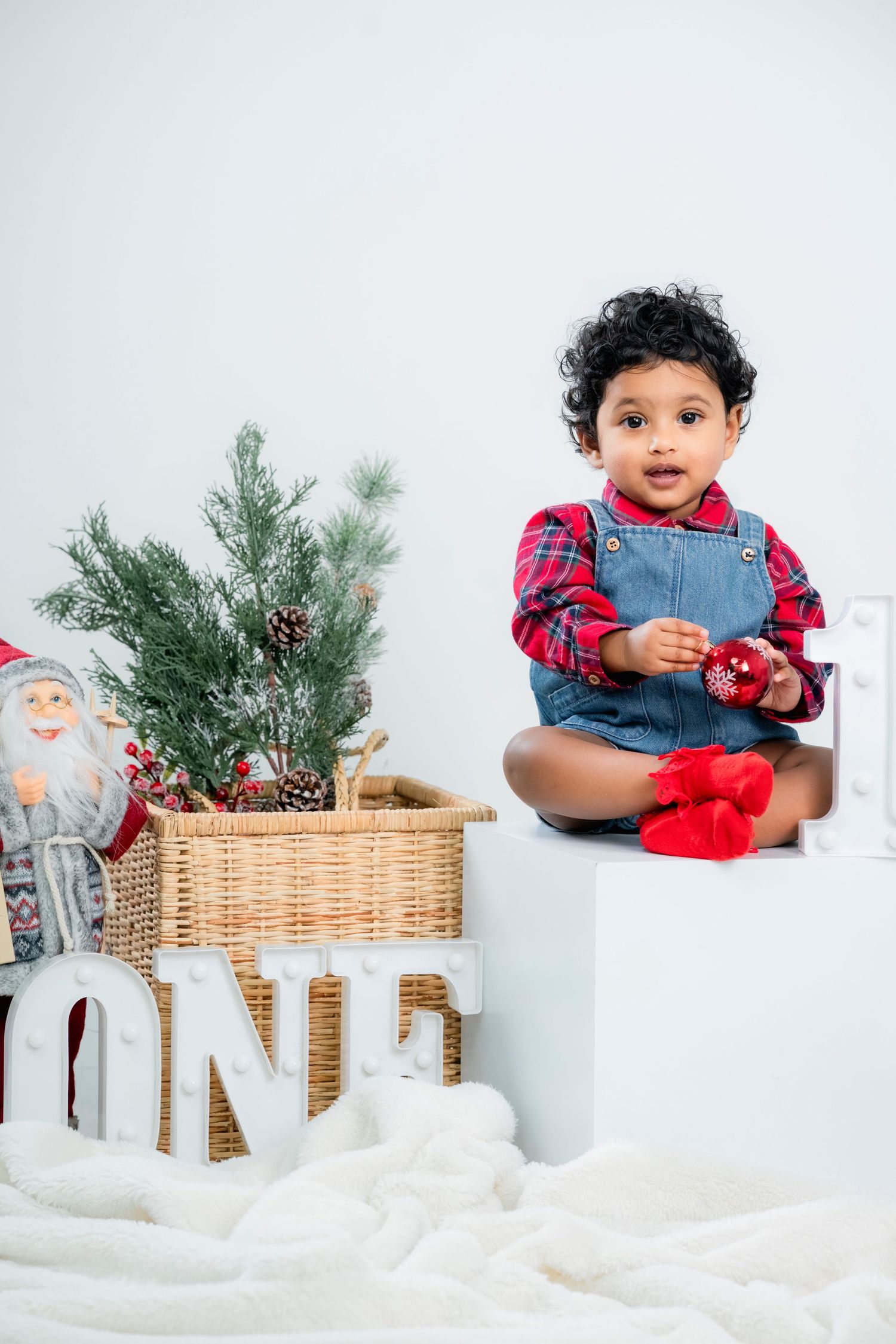 Holiday themed photo session with wicker basket, pine branches, and festive red and plaid decor against white backdrop.