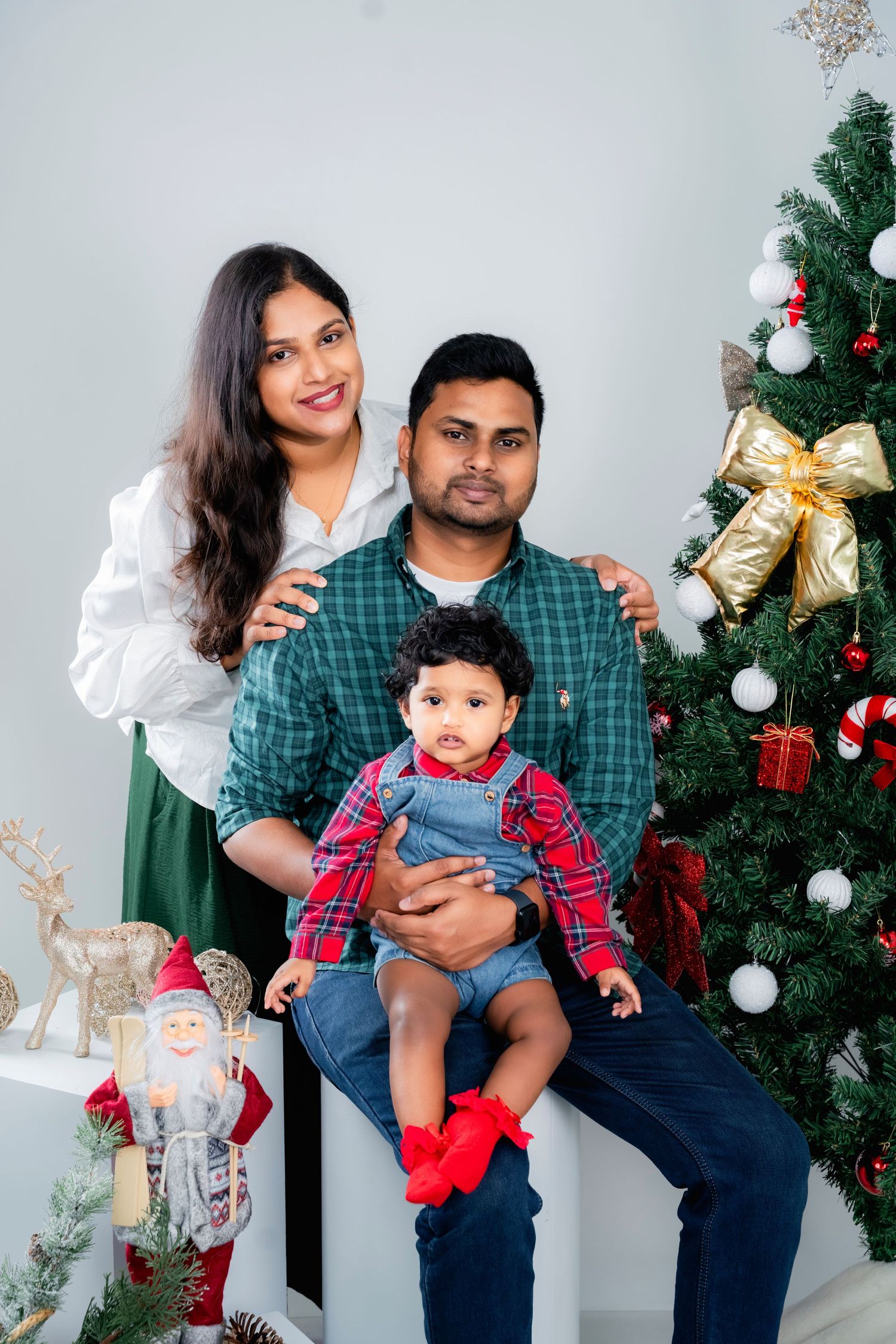 Family poses together in front of decorated Christmas tree wearing coordinated plaid and denim outfits.