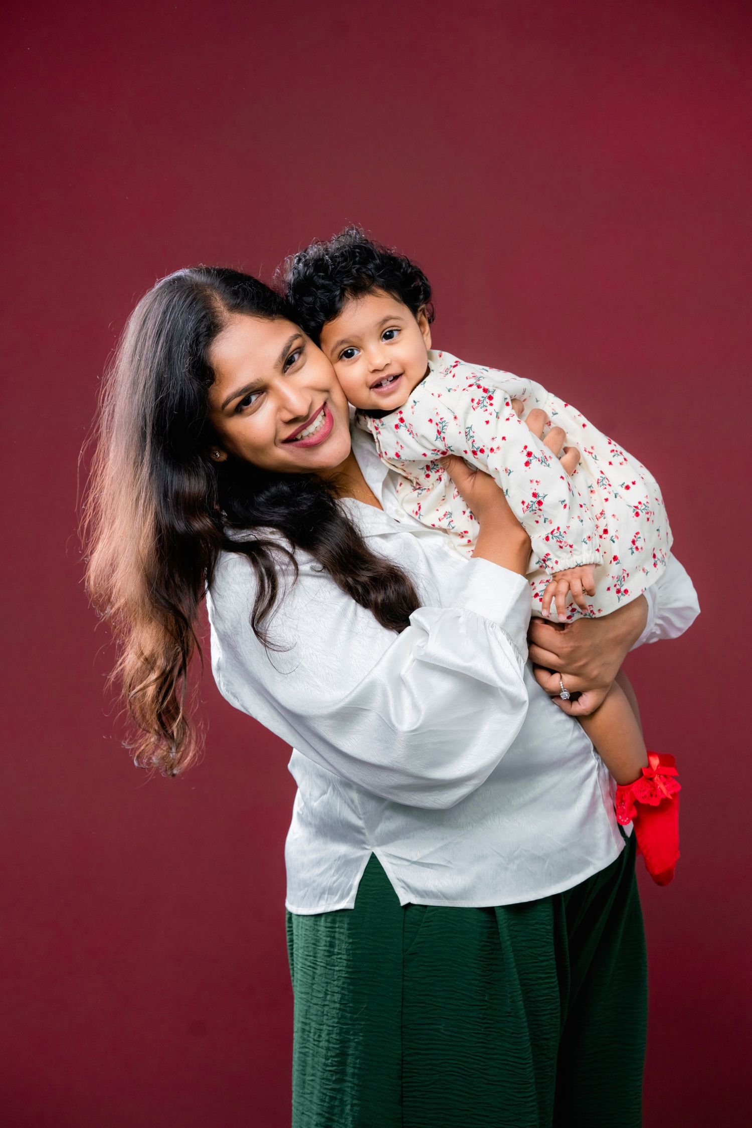 A mother in a white top and green skirt shares playful moments with her child against a burgundy background.