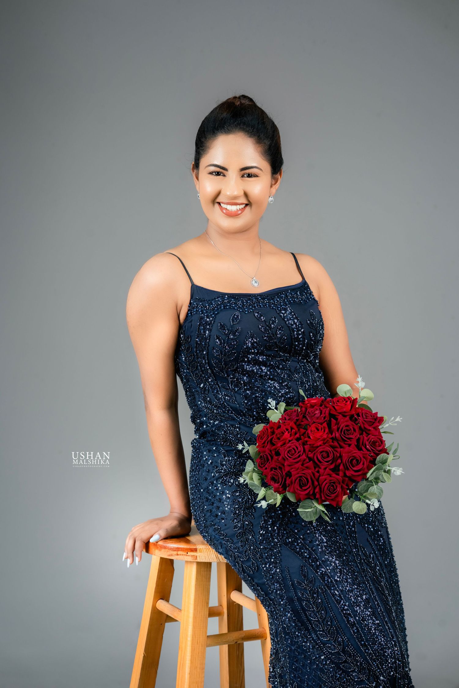 A person in a sparkling navy blue sequin dress poses with red roses on a wooden stool against a grey backdrop.