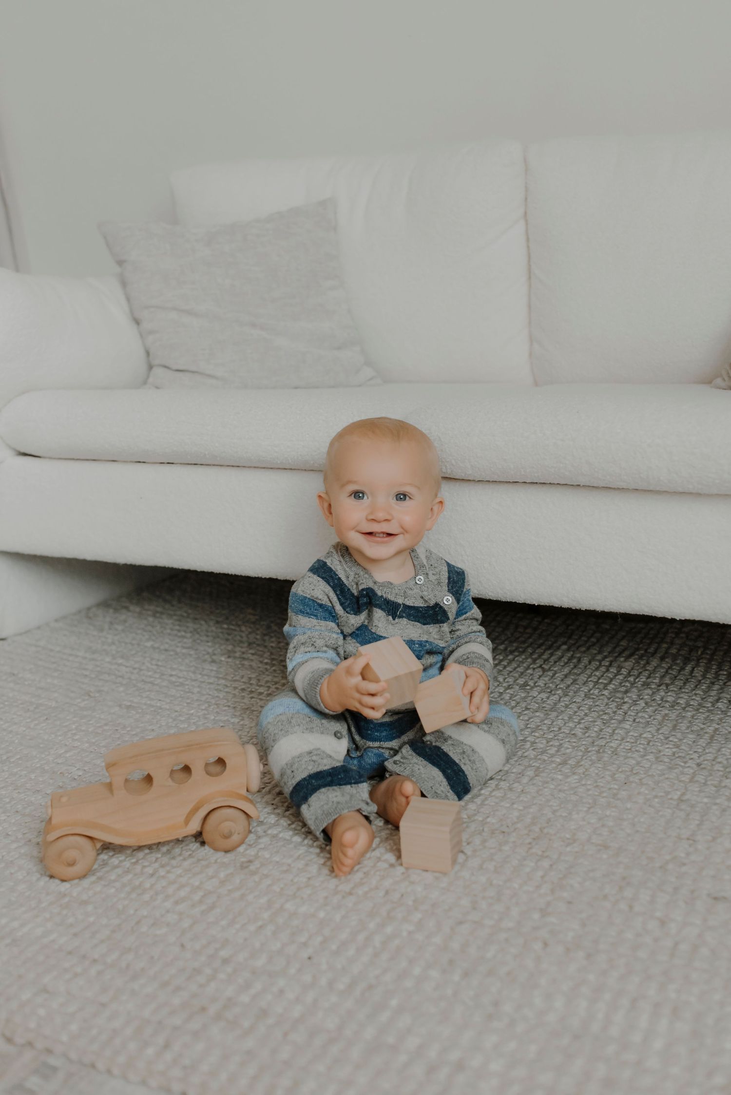 A small child in striped overalls sits on a gray carpet next to a wooden toy car beside a white couch.