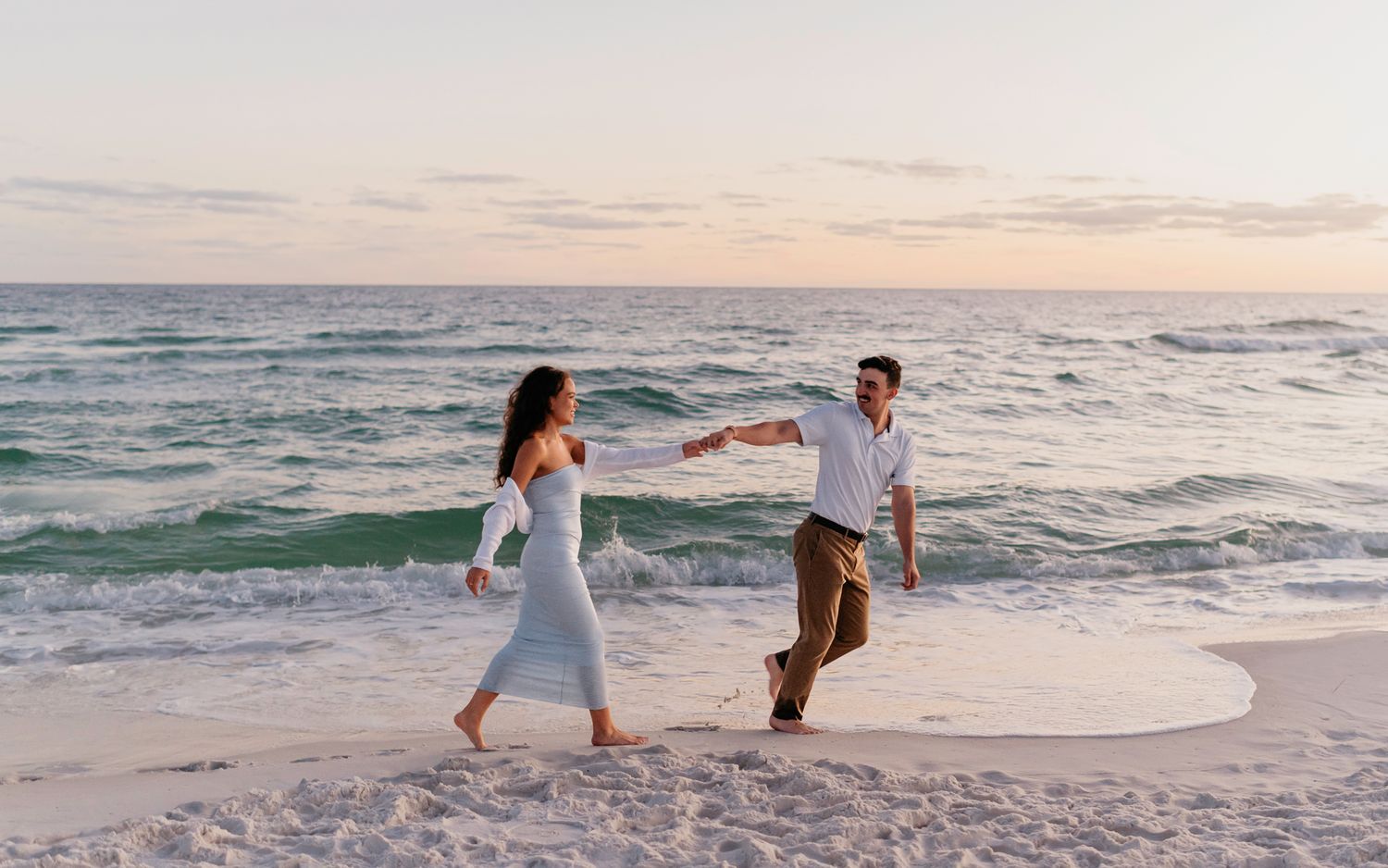A couple walks hand in hand along a sandy beach at sunset with waves crashing behind them.