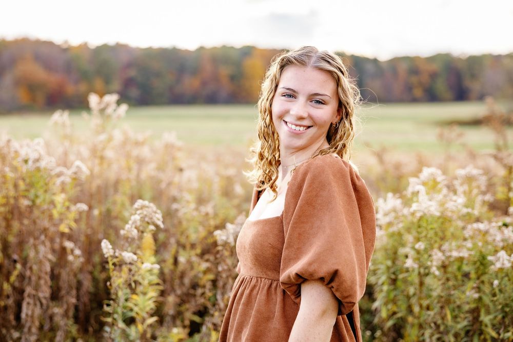 Senior in a brown dress stands in a sunlit autumn field with wildflowers and fall foliage in the background.