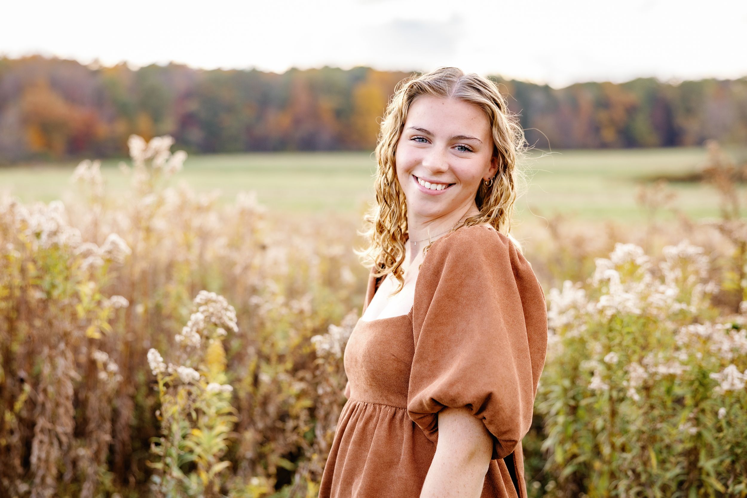 Senior in a brown dress stands in a sunlit autumn field with wildflowers and fall foliage in the background.