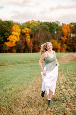 Senior in sage dress running joyfully through autumn field at sunset.