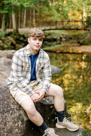 Senior sitting on rock by stream wearing plaid shirt and khaki shorts.