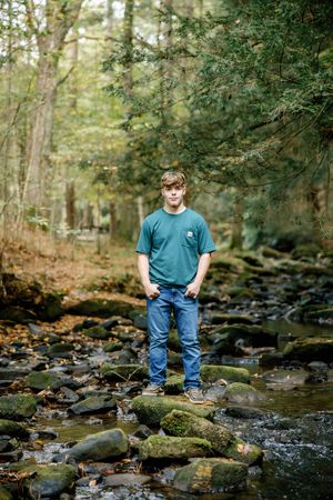 Senior in blue shirt and jeans standing in rocky forest stream.