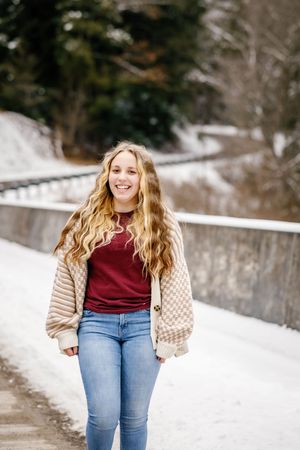 Senior in burgundy sweater and beige cardigan walking on snowy winter road.