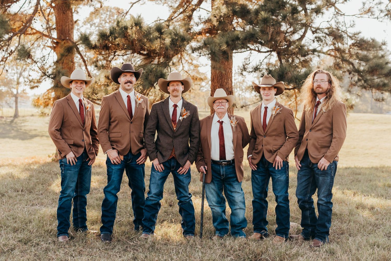 Wedding party members in brown jackets and blue jeans pose together outdoors during a rustic fall celebration.