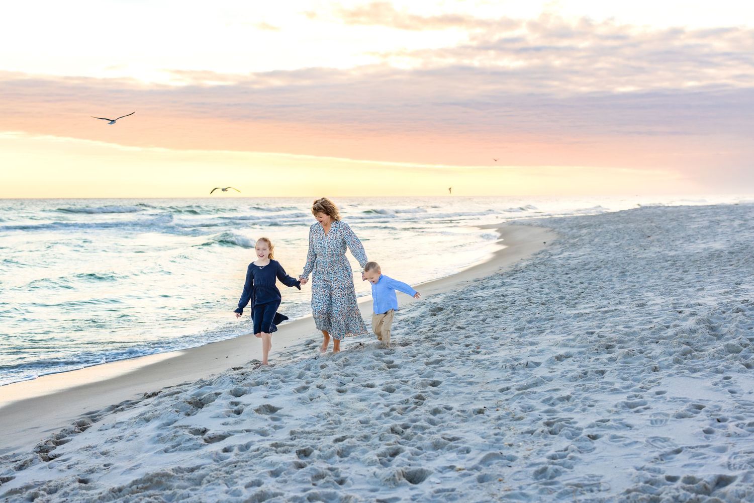 A parent and child walk along the ocean shore at sunset, leaving footprints in the sand as waves roll in.