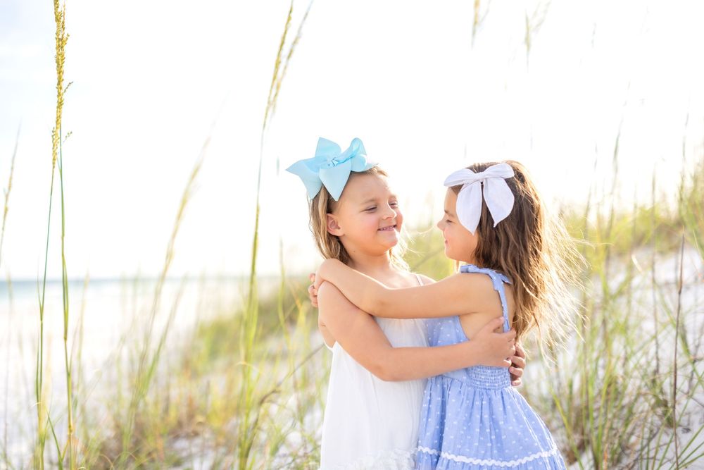 Two people in light blue dresses walk together on a sandy beach path during sunset.