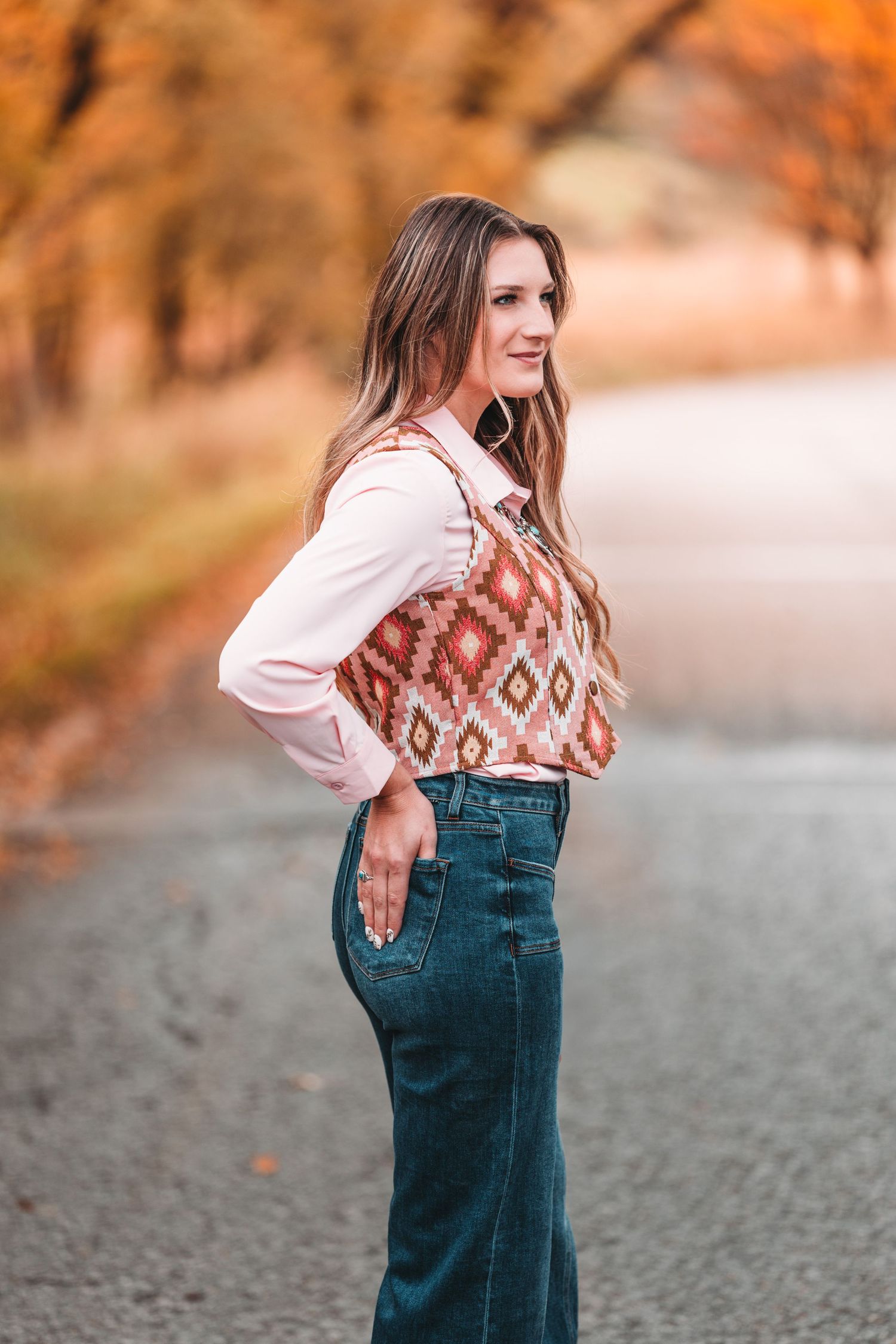A person in overalls and a patterned sweater poses playfully against an autumn backdrop with warm orange foliage.
