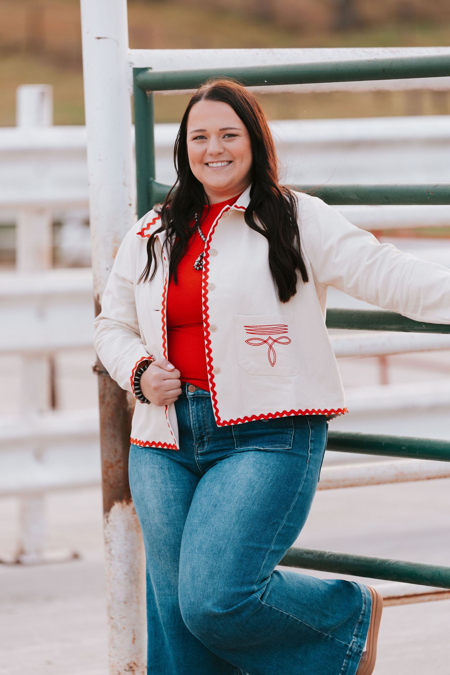 A person wearing a white and red embroidered jacket with blue jeans poses casually against a metal railing outdoors.