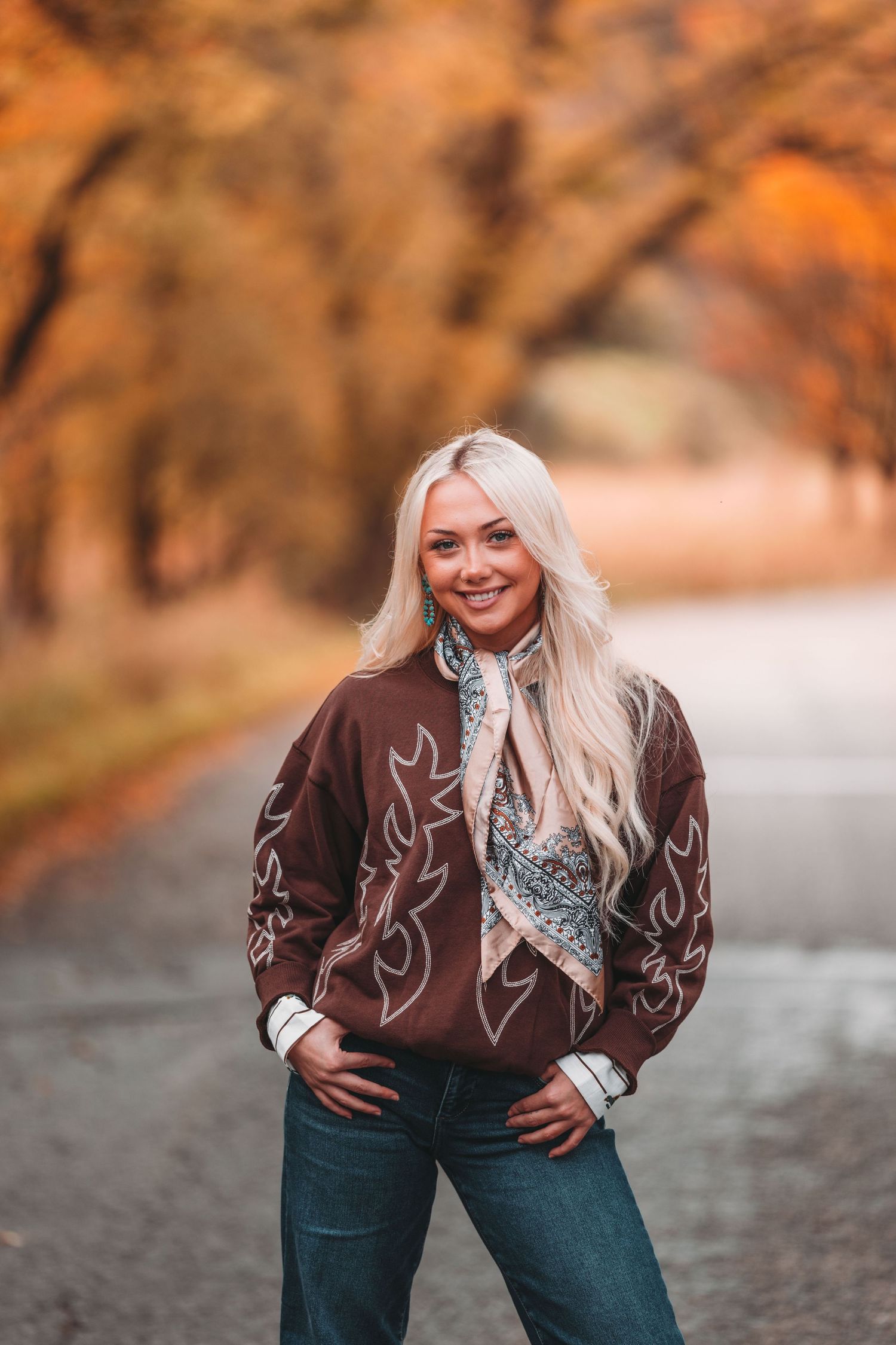 A person wearing a brown embroidered western shirt and jeans poses outdoors during autumn with warm lighting.