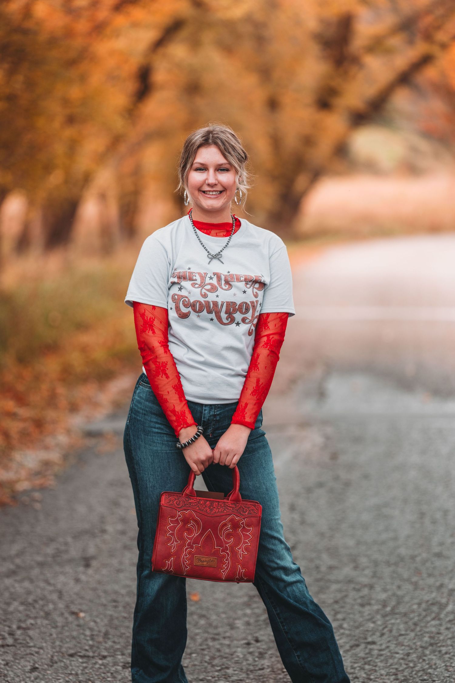 Person in casual western style outfit with red turtleneck, graphic t-shirt, jeans and red handbag on autumn path.