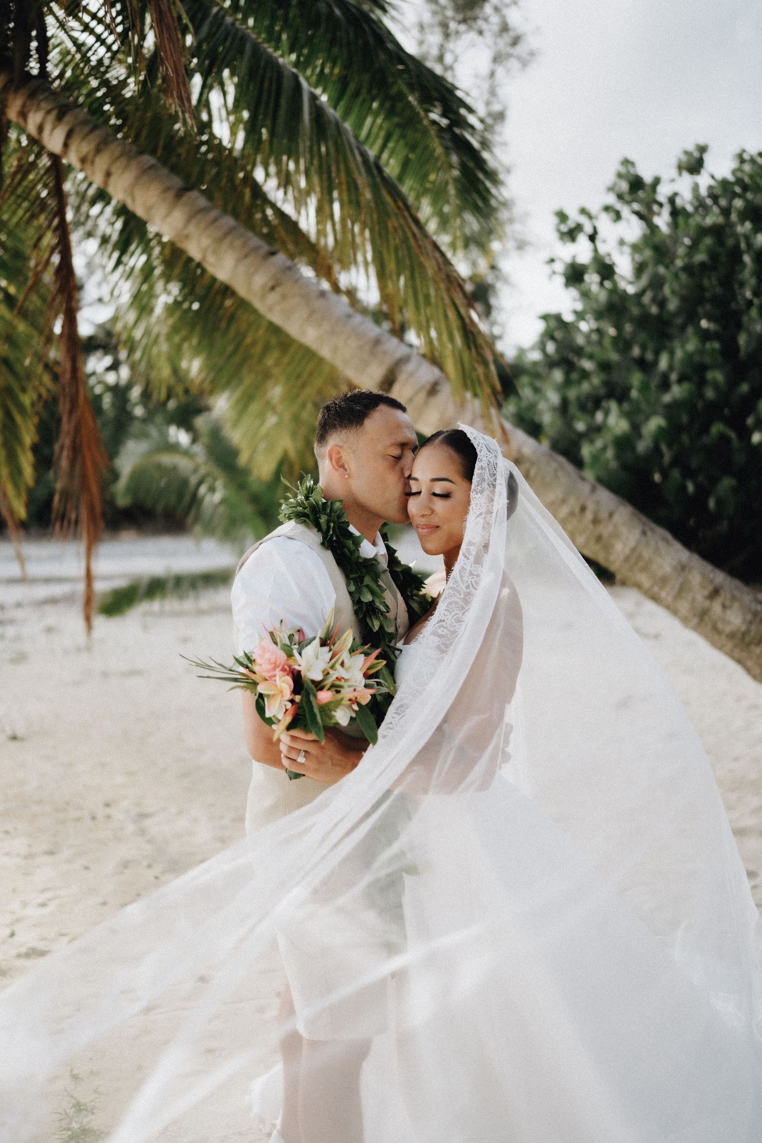 Newlyweds share a romantic kiss under palm trees on a tropical beach at sunset.