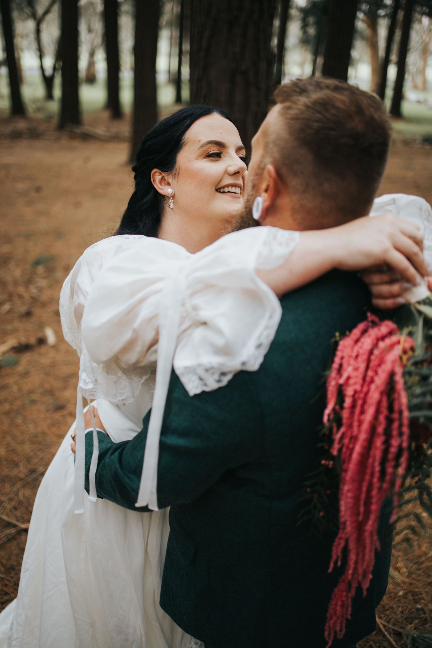 Joyful dark-haired bride in white puff-sleeve gown embraces groom in pine forest wedding portrait.