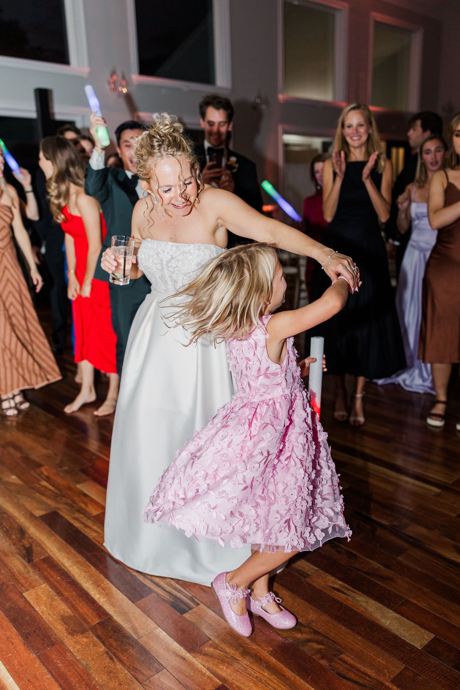 Wedding guests in colorful dresses dance and twirl on a hardwood floor under chandeliers.