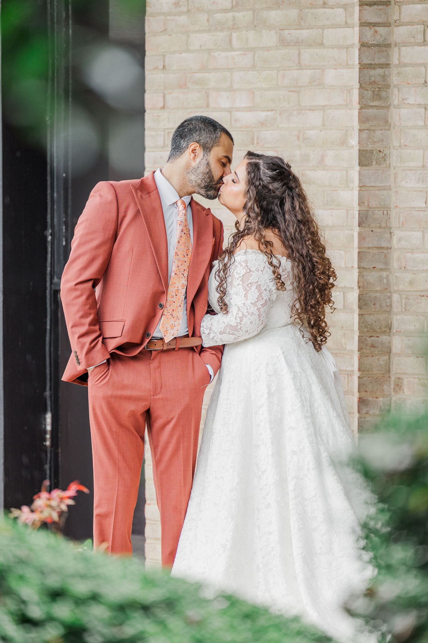 A romantic wedding couple in a coral suit and white lace dress share tender moments against a light brick wall.