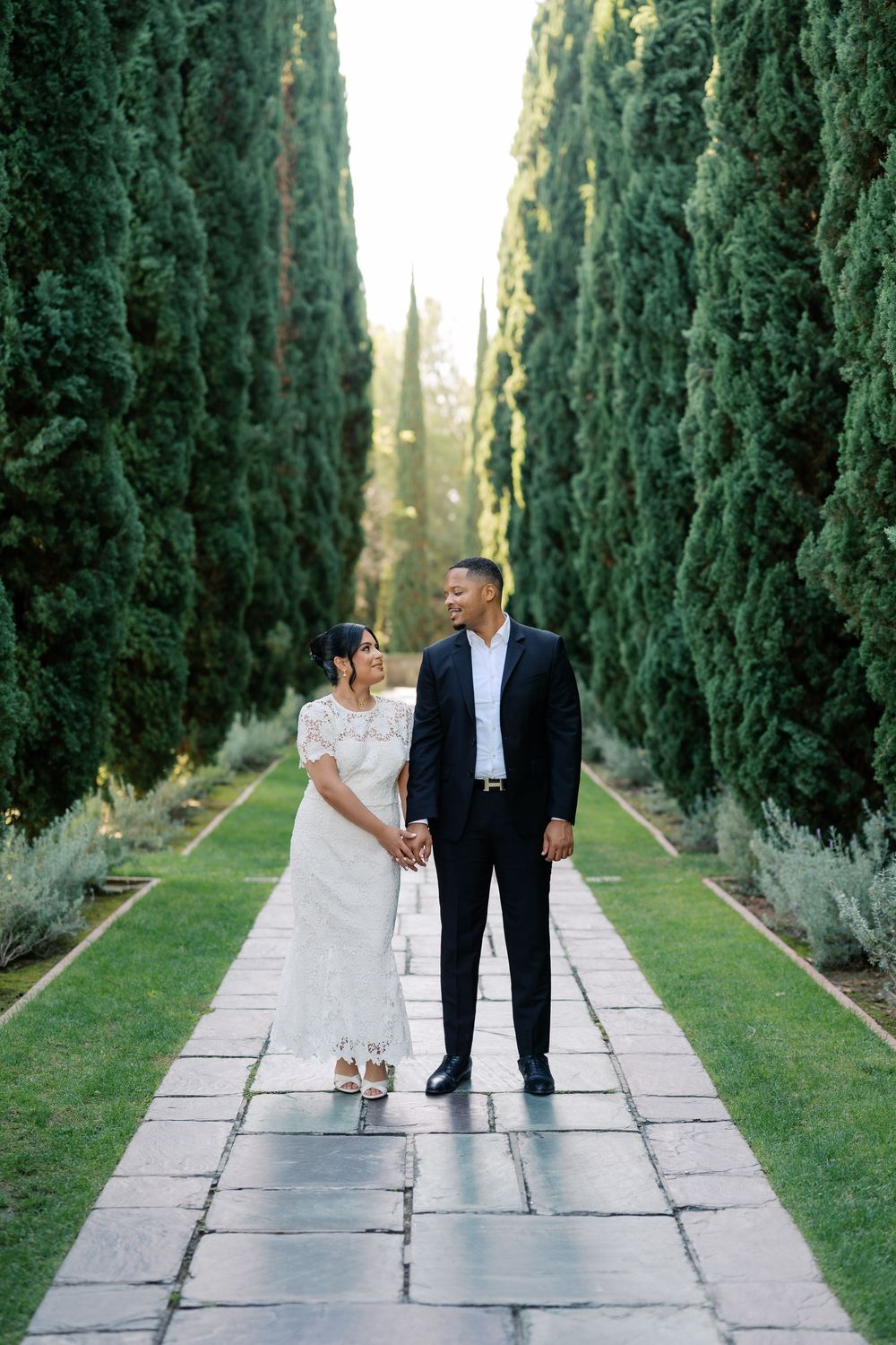 A couple walks together down a scenic cypress tree-lined path in formal attire on a sunny day.