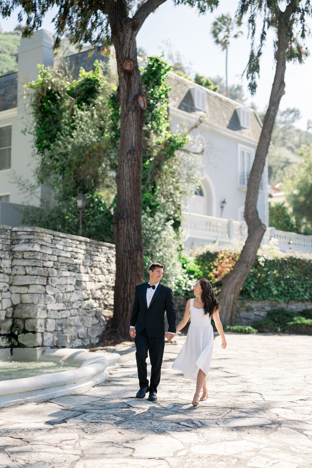 A couple in formal attire walks hand in hand along a stone path by a white Mediterranean-style building with trees.