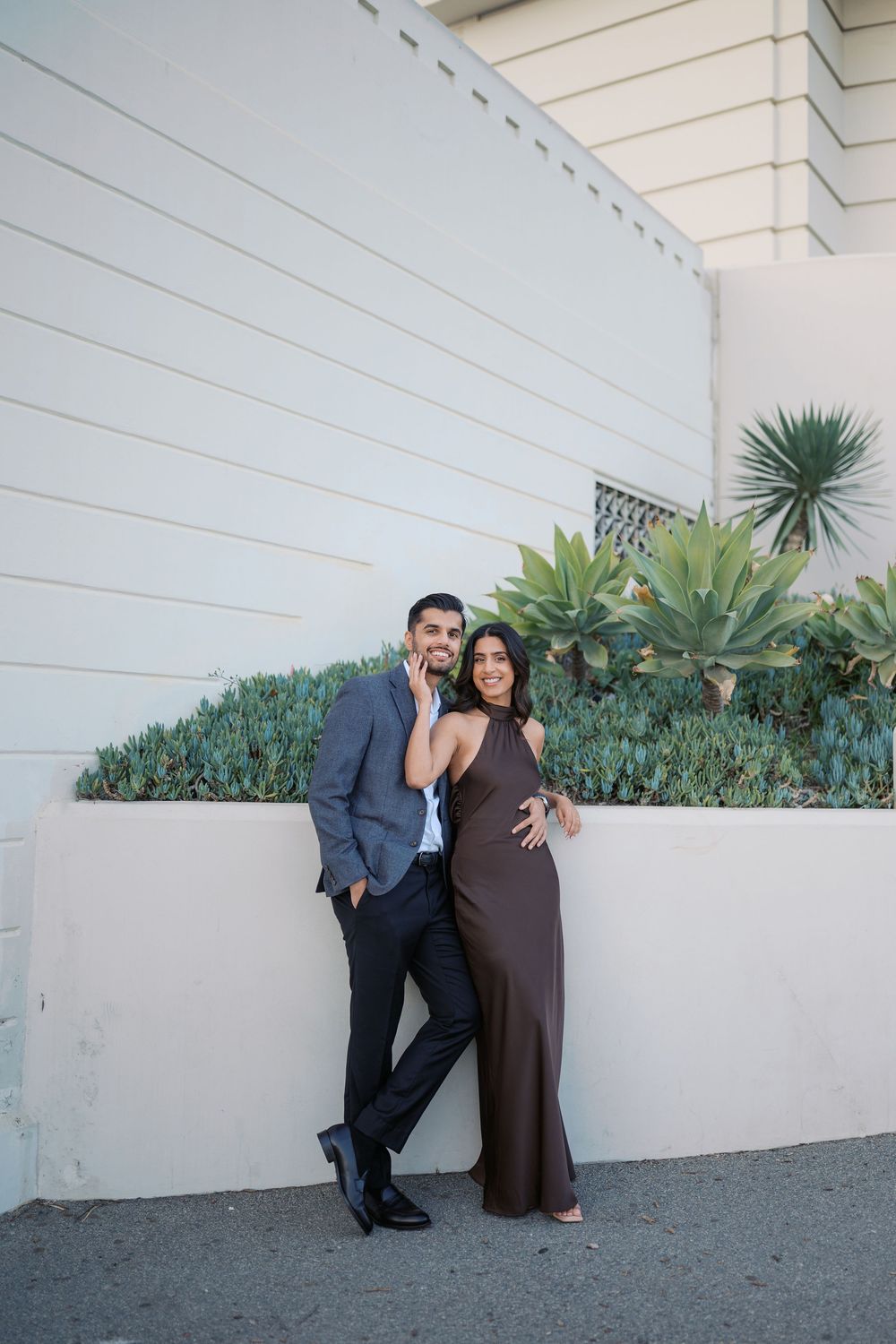 A couple poses together against a white wall with succulent plants in an outdoor setting.