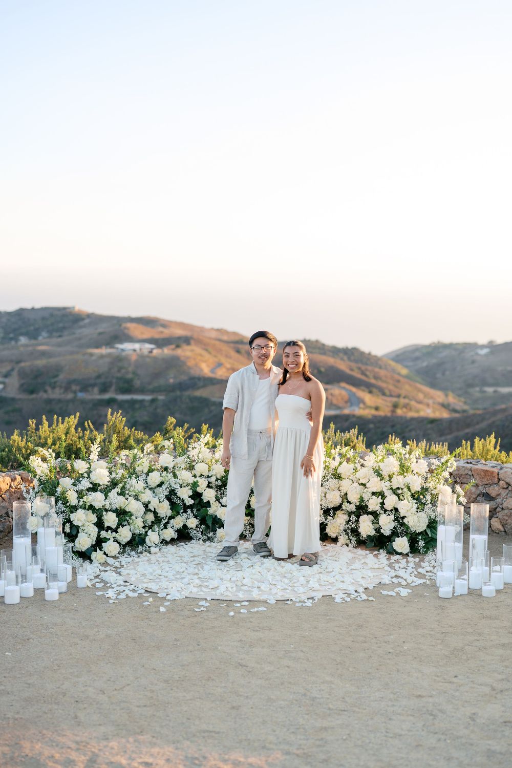 Elegant outdoor wedding ceremony setup with white floral arrangements and candles lining a sandy beach at sunset.