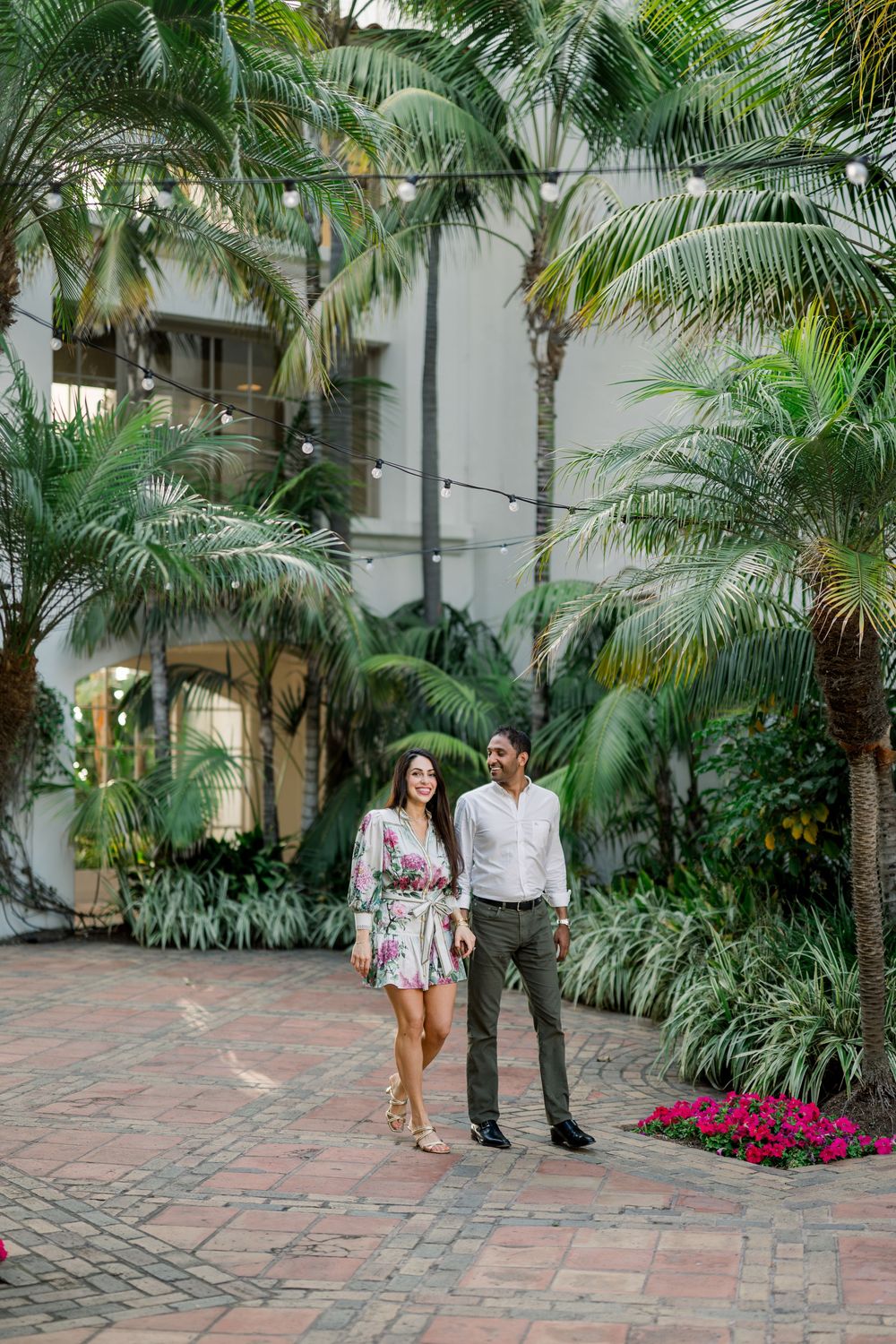Couple walking together on brick pathway surrounded by lush tropical palm trees and pink flowers in garden setting.
