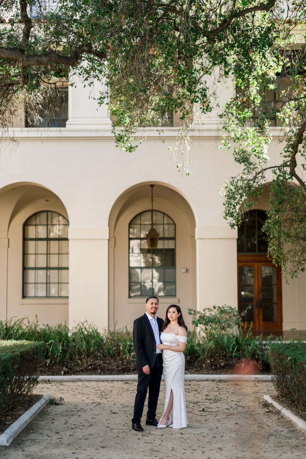 A couple shares a romantic moment under green trees at an elegant arched building with white columns.