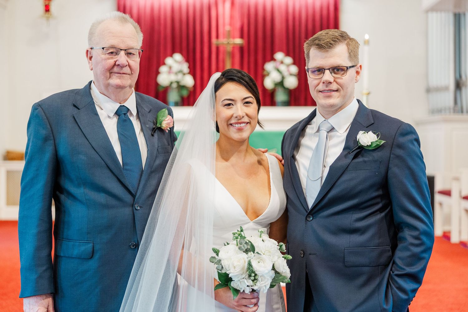 Three people in formal attire pose together at a church wedding ceremony with red carpet and white decorations.