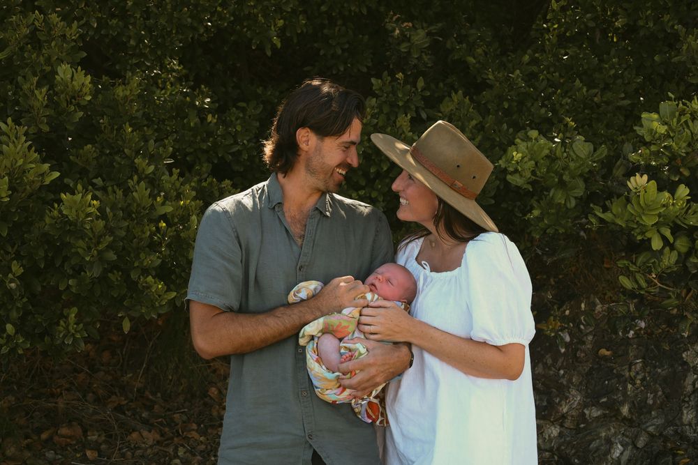 A couple holding a newborn baby stands together in a garden setting surrounded by lush greenery.