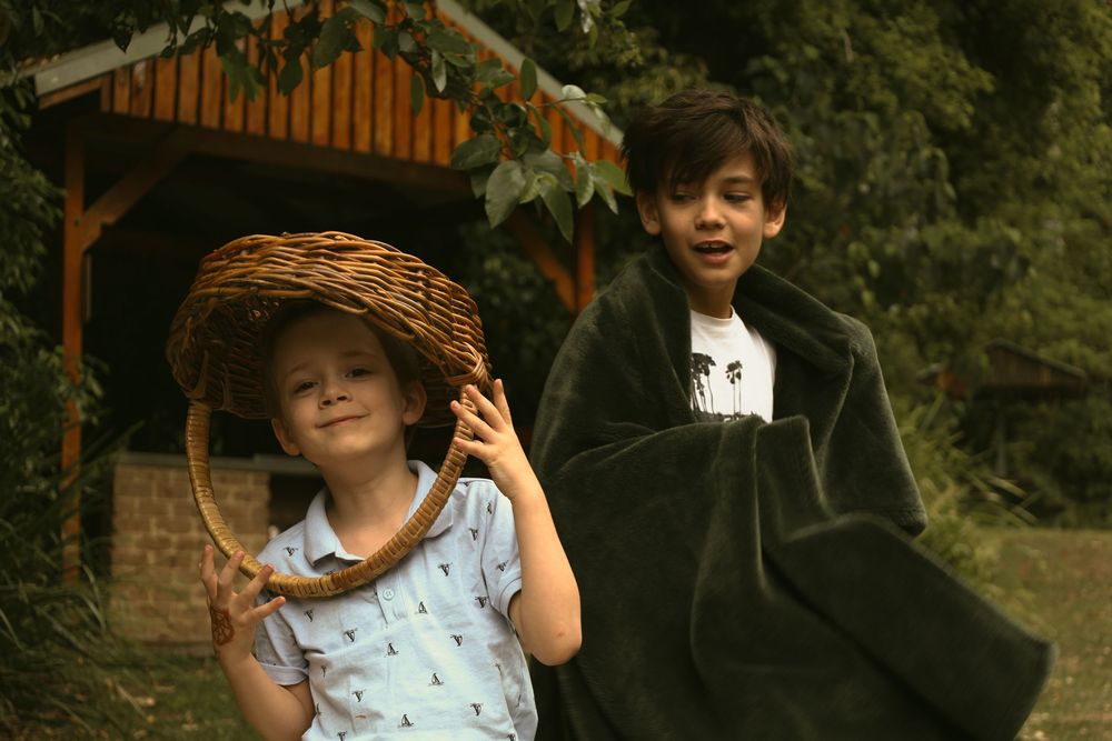 Two people pose playfully outdoors near a wooden gazebo, one wearing a straw hat and summer dress, the other in dark clothing.
